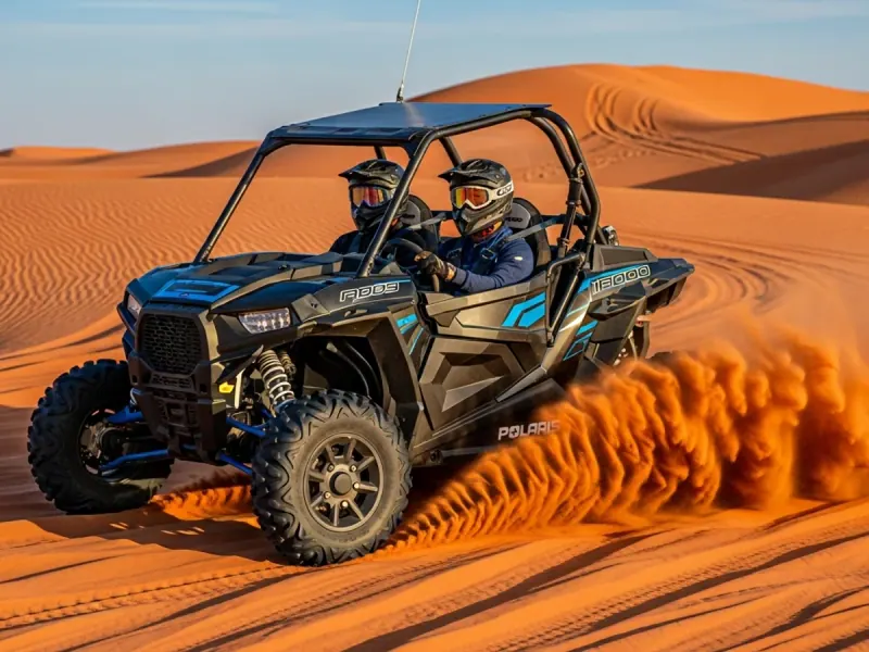 Two helmeted riders in a black Polaris RZR throw up a plume of orange sand.